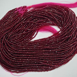 May include: A large bundle of small, faceted, deep red beads. The beads are strung together, creating multiple strands. A bright pink tassel is attached to the top of the bundle. The beads have a sparkling, reflective quality.