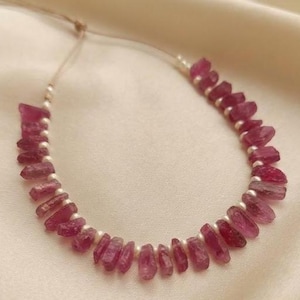 Puede incluir: Un collar que presenta una serie de fragmentos de piedras preciosas en bruto de color rosa intenso que se alternan con pequeñas cuentas blancas redondas. Las piedras son translúcidas y de forma irregular, ensartadas en un cordón fino claro.