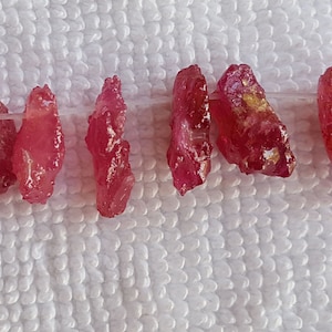 Puede incluir: Una hebra de cuentas de piedras preciosas de color rojo rubí, cada cuenta tiene una forma irregular y tosca. Las cuentas están enhebradas en un hilo transparente y están dispuestas en línea.