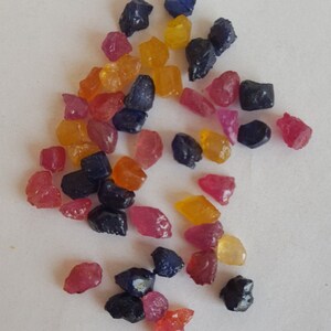 Puede incluir: Una colección de piedras preciosas sueltas en varios colores, incluyendo azul, rojo y amarillo. Las piedras preciosas son ásperas y sin pulir, y están esparcidas sobre una superficie blanca.