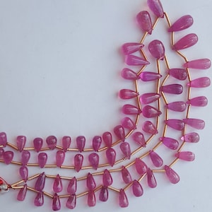 May include: A strand of pink gemstone beads in a teardrop shape. The beads are strung on gold wire.