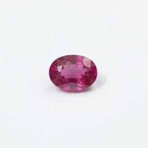 May include: A vibrant, oval-cut gemstone with a deep magenta-pink hue. The stone features a faceted surface that reflects light, creating a brilliant, sparkling appearance across its polished, symmetrical shape against a plain white background.