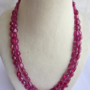 Könnte beinhalten: Eine zweireihige Halskette mit polierten, ovalen Rubinperlen. Die Perlen haben eine tiefe, lebendige rosa-rote Farbe und sind so aufgereiht, dass ein mehrschichtiger Effekt entsteht. Die Halskette wird auf einer weißen Büste präsentiert, die das Design des Schmucks hervorhebt.