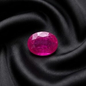 May include: A vibrant, oval-cut gemstone in a deep, saturated magenta-pink hue. The stone features a faceted surface that reflects light, showcasing a brilliant, polished finish with clear internal clarity and a distinct, symmetrical oval shape.
