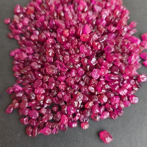 May include: A collection of raw ruby gemstones, displaying a deep red hue. The uncut stones have a slightly translucent quality and vary in size and shape. The pile of gems creates a natural, textured appearance.