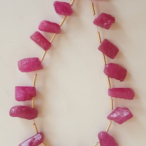 May include: A strand of pink gemstone beads strung on a gold wire. The beads are irregular in shape and have a rough, natural texture.