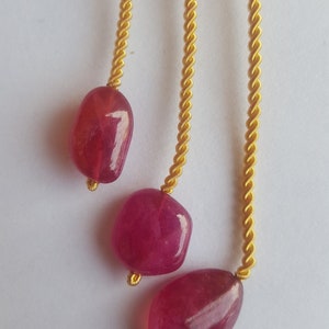 May include: Three pink gemstone beads hang from a gold cord with a woven fabric top.