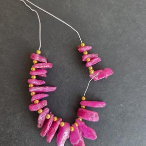 Puede incluir: Un collar con cuentas de piedras preciosas de color rosa vibrante, de forma rugosa e irregular. Las cuentas están ensartadas en un fino alambre plateado e intercaladas con pequeñas cuentas doradas. El collar se muestra sobre un fondo oscuro.