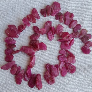 Puede incluir: Una hebra de cuentas de rubí rosa, cada cuenta tiene una forma irregular y tosca. Las cuentas están enhebradas en un hilo transparente y están dispuestas en un círculo suelto.