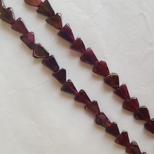 May include: Two strands of dark red, triangular beads. The beads are strung on a red thread and are arranged diagonally across a white background. The beads have a translucent quality.
