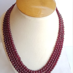 May include: A three-strand necklace made of dark red beads. The necklace is displayed on a white mannequin bust.
