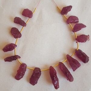 May include: A strand of 14 raw, unpolished ruby gemstone beads, strung on a gold wire. The beads are a deep, rich red color and have a natural, irregular shape.
