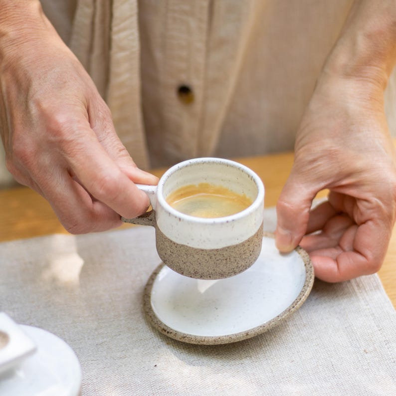 May include: A small ceramic espresso cup and matching saucer featuring a two-tone design. The upper portion of the cup and the center of the saucer are glazed in glossy white, while the lower portion and outer rim have a speckled brown finish.