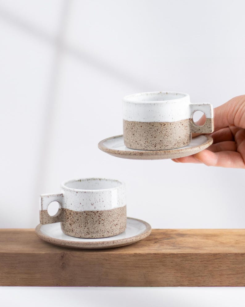 May include: Two ceramic espresso cups with matching saucers, each featuring a two-tone design with a speckled brown base and a smooth white upper section. The cups have a distinct square-shaped handle with a circular cutout in the center.