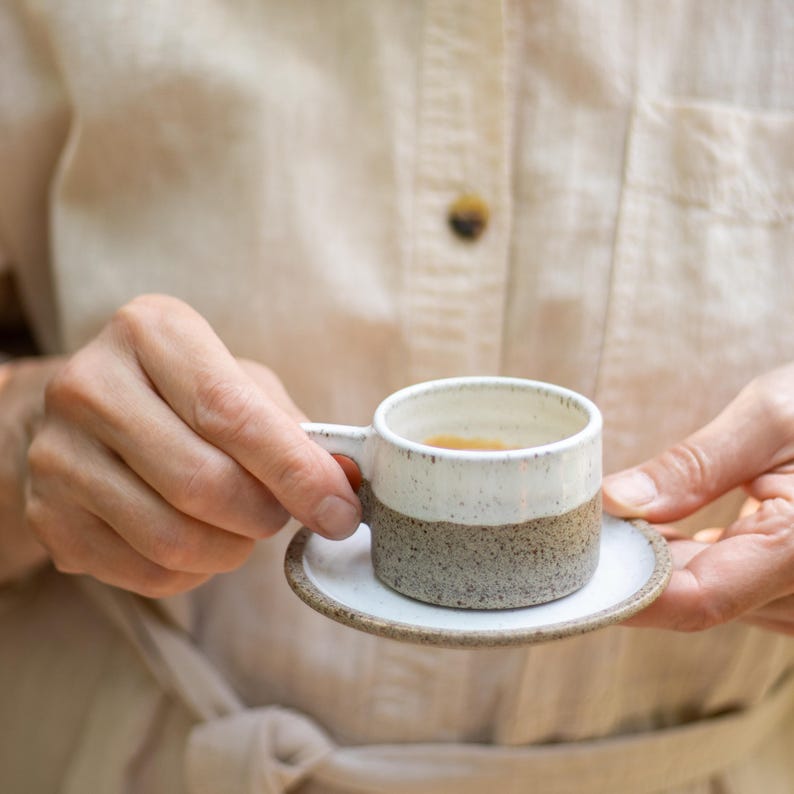 May include: A small ceramic espresso cup and matching saucer featuring a two-tone design. The upper portion of the cup is glazed in a smooth white, while the lower half and the saucer display a speckled, textured grey and brown earth-toned finish.