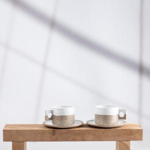 May include: Two ceramic espresso cups with matching saucers, each featuring a speckled tan base and a smooth white upper section. The cups have a distinct square-shaped handle with a circular cutout, showcasing a two-tone design and texture.