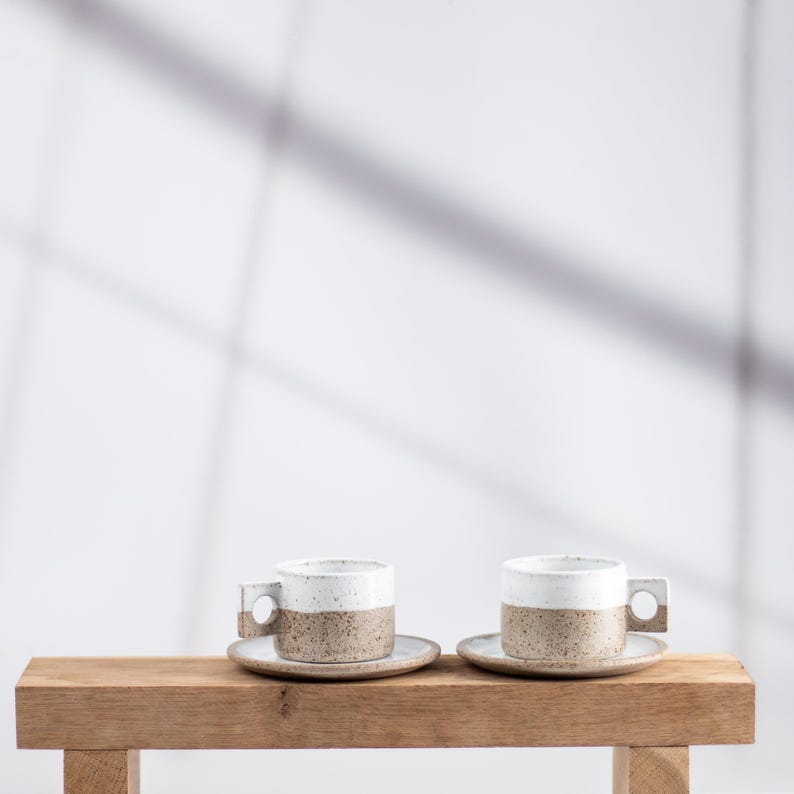 May include: Two ceramic espresso cups with matching saucers, each featuring a speckled tan base and a smooth white upper section. The cups have a distinct square-shaped handle with a circular cutout, showcasing a two-tone design and texture.