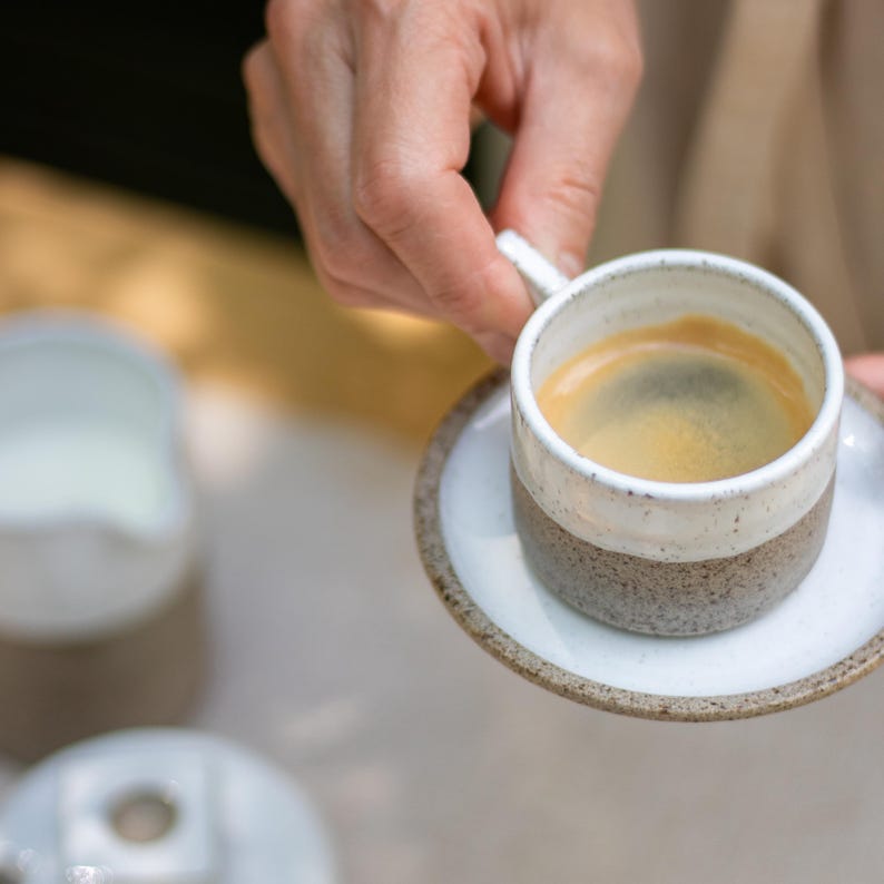 May include: A ceramic espresso cup and matching saucer featuring a two-tone design. The upper portion of the cup and the center of the saucer are glazed in white, while the lower portion of the cup and the saucer rim have a speckled brown finish.