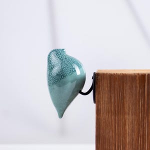 Könnte beinhalten: Eine türkisfarbene Keramikvogel-Figur mit einer gesprenkelten, rissigen Glasuroberfläche. Der Vogel ist an einem schwarzen Metallhaken befestigt, der an der Seite eines rechteckigen Blocks aus natürlichem hellbraunem Holz mit sichtbarer Maserung montiert ist.