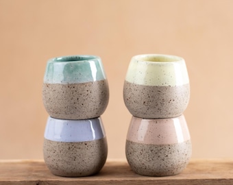 May include: Four small ceramic cups arranged in two stacks of two. Each cup features a speckled brown base and a smooth, solid-colored upper section in light blue, lavender, pale yellow, and soft pink, with a rounded, handleless design.