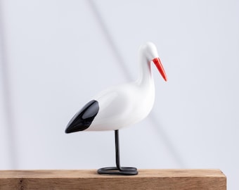May include: A stylized bird figurine featuring a smooth white body, a bright orange beak, and black wing tips. The bird stands on a thin, black metal wire leg that forms a circular base, creating a clean and simple representation of a stork.