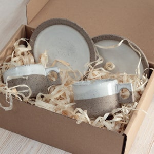 May include: A set of two ceramic mugs and two matching saucers nestled in a cardboard box with wood shavings. Each mug features a white glazed upper section and a speckled grey textured lower section with a square handle containing a circular cutout.
