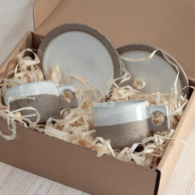 May include: A set of two ceramic mugs and two matching saucers nestled in a cardboard box with wood shavings. Each mug features a white glazed upper section and a speckled grey textured lower section with a square handle containing a circular cutout.