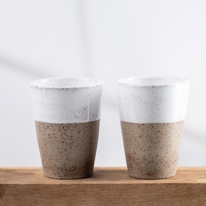 Pode incluir: Dois copos de cerâmica com um design de dois tons. A parte superior tem um acabamento de esmalte branco brilhante com pequenas manchas castanhas, enquanto a parte inferior consiste numa textura de argila bege sem esmalte com manchas escuras.