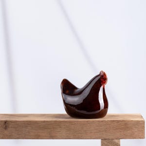 Puede incluir: Una figura de gallina de cerámica de color marrón oscuro brillante con una superficie lisa y reflectante. La escultura presenta un detalle distintivo de cresta y barbillas rojas en la cabeza, capturando la forma redondeada de una gallina sentada.
