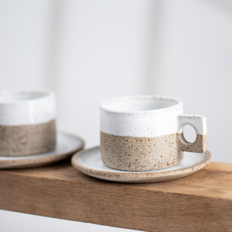 May include: Two ceramic cups and matching saucers featuring a two-tone design. The upper portion of each cup is smooth white, while the lower section and the saucer are a speckled, textured tan clay. Each cup has a square handle with a circular cutout.