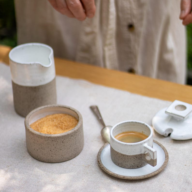 May include: A ceramic coffee set featuring a tall pitcher, a small bowl filled with brown sugar, and a cup with a handle on a matching saucer. Each piece has a speckled beige base and a smooth white glazed top. A silver spoon lies nearby.