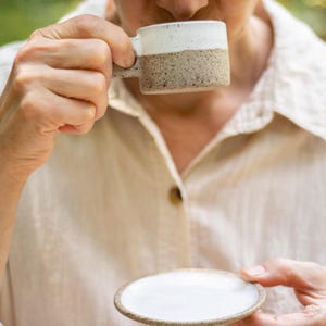 May include: A small ceramic mug and matching saucer featuring a two-tone design. The upper portion of the mug and the center of the saucer are glazed in smooth white, while the lower portion of the mug and the saucer rim have a speckled tan finish.