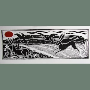 May include: Black and white linocut print of a red sun, two birds flying, and three rabbits in a field. The print is framed in a black border.
