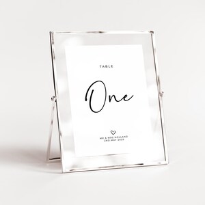 May include: A silver table number holder with a white card that says "Table One" in black script. The card also includes the text "Mr & Mrs Holland 2nd May 2023" and a small heart symbol.