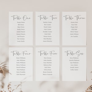 May include: Six white cards with black text listing table numbers and guest names. The cards are arranged in two rows of three. The cards are on a light beige background.