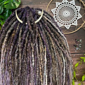 May include: Close-up of a set of dreadlocks in shades of purple, gray, and brown. A silver-toned hair accessory with a pearl accent is pinned into the dreads. A white, lacy dreamcatcher is in the background.
