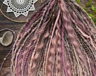 May include: A bundle of handmade dreadlocks in shades of pink, gray, and brown. The dreads have a mix of textures, including wavy and braided sections. A dreamcatcher and other decorative items are in the background.