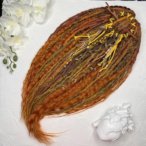 Puede incluir: Un conjunto de rastas hechas a mano en tonos naranja, marrón y verde. Las rastas están decoradas con cintas amarillas, pequeñas cuentas y vegetación artificial. El cabello está peinado con un adorno decorativo en forma de cuerno.
