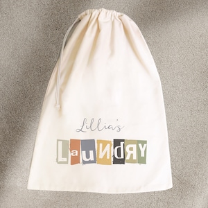 May include: A white canvas laundry bag with a drawstring closure. The bag has the text "Lillia's Laundry" printed on it in colorful, block letters.