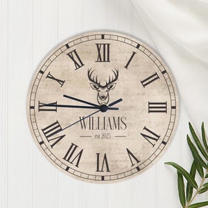 May include: A round wall clock with a vintage design. The clock face is beige with black Roman numerals and hands. A deer head graphic is centered above the name "WILLIAMS" and the year "est 2025".
