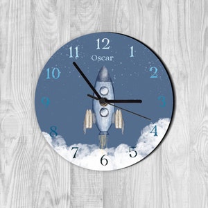 May include: A blue and white rocket ship clock with the name "Oscar" on the face. The clock has a white cloud background with blue numbers.