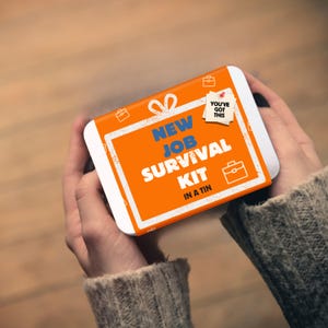 May include: A rectangular metal tin featuring an orange label with a white gift bow graphic. The text reads "NEW JOB SURVIVAL KIT IN A TIN" in blue and white, with a small white note pinned to the top right corner that says "YOU'VE GOT THIS".