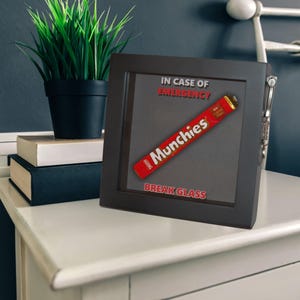May include: A black framed box with a glass front. The box has a red chocolate bar inside with the text "Munchies" on it. The text "In case of emergency, break glass" is printed on the box.