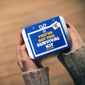 Könnte beinhalten: Eine rechteckige blaue Dose mit weißem Rand wird mit beiden Händen gehalten. Auf der Dose steht "YOU'VE GOT THIS SURVIVAL KIT IN A TIN" in weißer Schrift. Die Dose hat auch ein weißes Etikett mit der Aufschrift "PACKED WITH FUN".