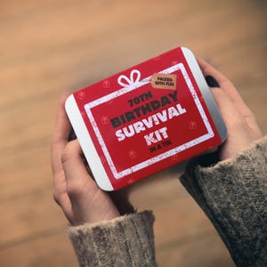 May include: A red tin with white text that reads "70th Birthday Survival Kit in a Tin." The tin has a white border and small gift box icons. A small tag reads "Packed with Fun."
