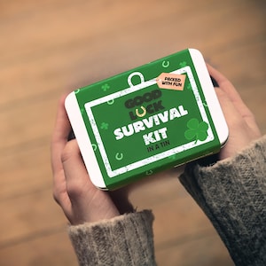 May include: A green and white rectangular tin with the words "Good Luck Survival Kit in a Tin." The tin has a horseshoe, shamrocks, and the phrase "Packed with Fun." The tin is held in two hands.
