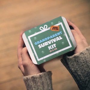 Puede incluir: Una lata rectangular verde con las palabras "Grandparent Survival Kit in a Tin" en blanco y azul. La lata tiene un borde blanco y está decorada con pequeños iconos de cajas de regalo. Una etiqueta naranja dice "Packed with Fun".