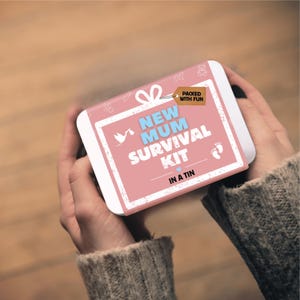 Puede incluir: Una lata de metal rectangular con una etiqueta rosa con borde blanco y el texto NEW MUM SURVIVAL KIT IN A TIN. La etiqueta incluye un icono de cigüeña blanca, un pequeño corazón azul, huellas de bebé y una etiqueta marrón que dice PACKED WITH FUN.