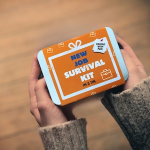 May include: A rectangular tin with a light blue border and an orange center, held in two hands. The tin reads "NEW JOB SURVIVAL KIT IN A TIN" in white and blue text, with a small tag that says "PACKED WITH FUN".