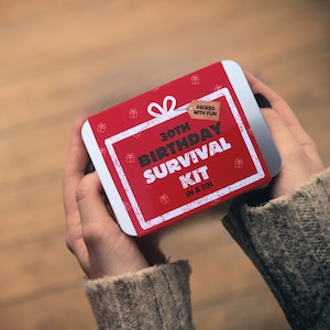 Könnte beinhalten: Eine rote, rechteckige Dose mit dem Text "30th Birthday Survival Kit" in Weiß. Die Dose wird in zwei Händen gehalten und hat ein kleines Etikett mit der Aufschrift "Packed with Fun". Die Dose ist mit kleinen Geschenkbox-Symbolen verziert.